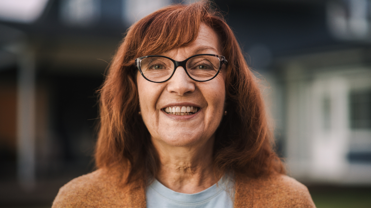 Close Up Portrait of a Cheerful Senior Woman with Ginger Hair Wearing Glasses Standing Outdoors in Front of a Residential Area Home. Retired Adult Female Looking at Camera and Smiling.