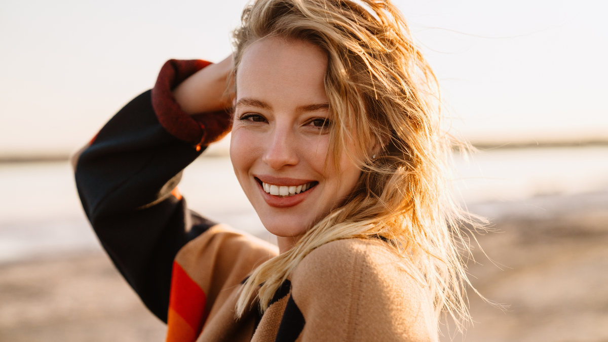 Happy young white woman smiling while walking by seashore on sunny day.