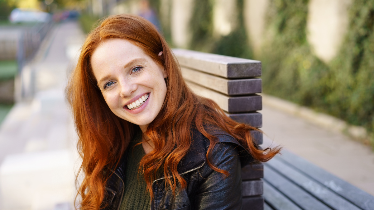 Attractive redhead woman relaxing on a bench outdoors turning to look at the camera with a happy friendly smile.