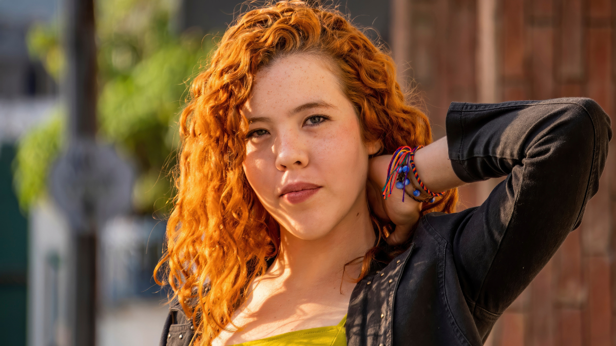 A woman with red hair poses for a picture. She is wearing a yellow dress and a black jacket.