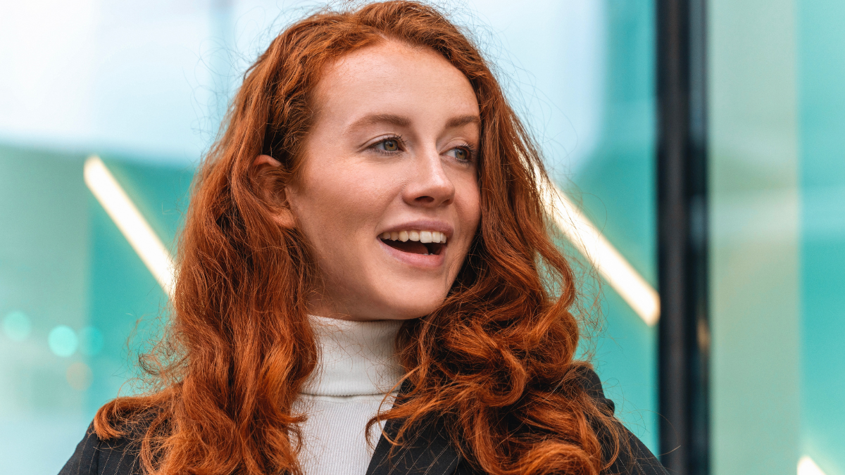 Woman with red hair smiling outside a modern building in urban setting during daytime.