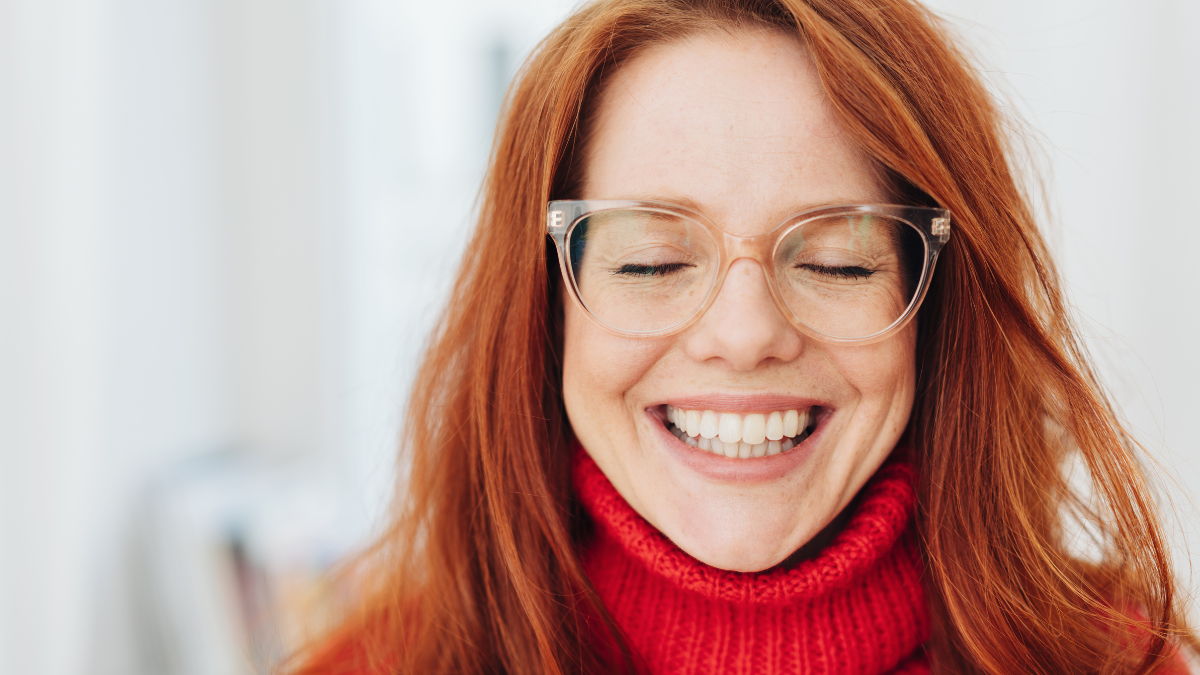 Pretty young woman wearing glasses beaming at the camera with a toothy grin as she closes her eyes.