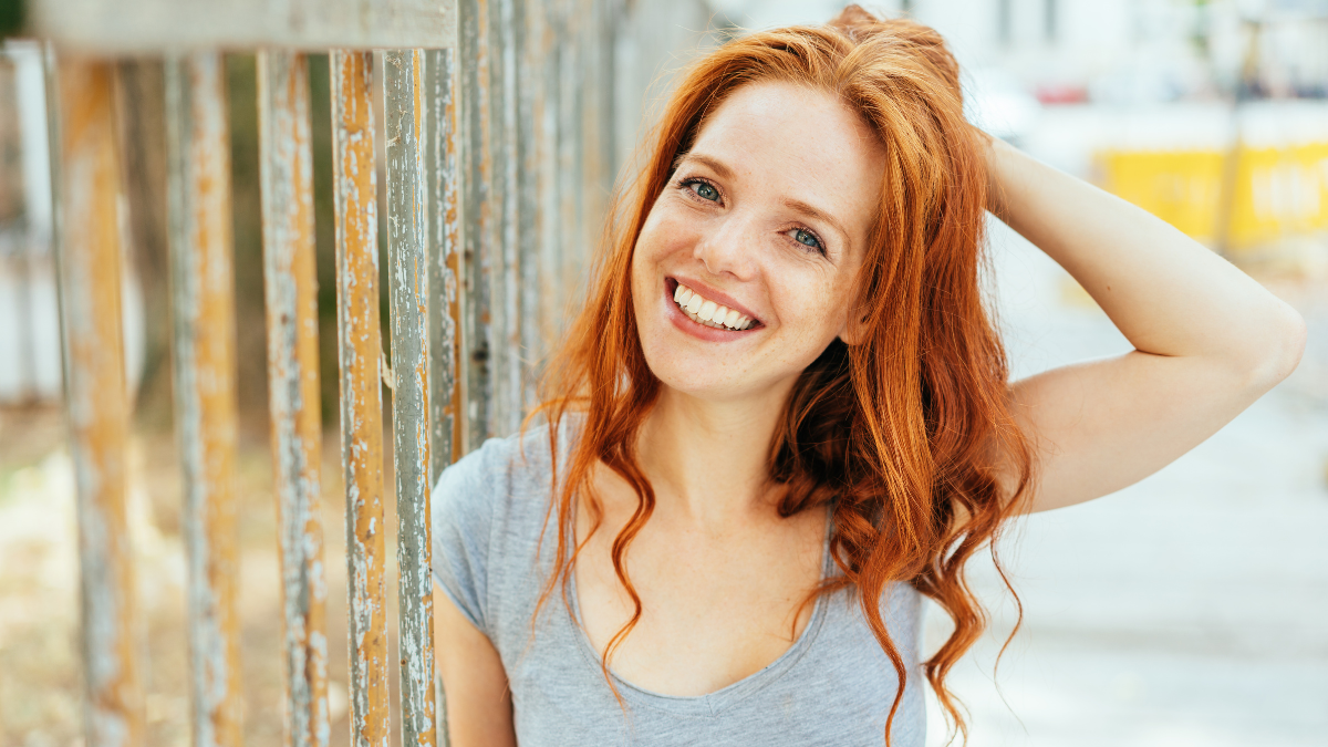 attractive friendly woman with tousled long red hair standing against a metal railing in a high key outdoor street.