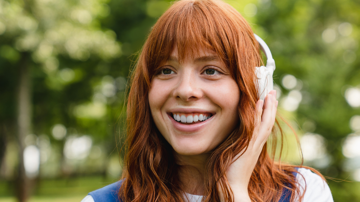Cheerful caucasian red-head young woman girl student teenager listening to the music in headphones, choosing sound track, playlist, favorite singer song outdoors in park.