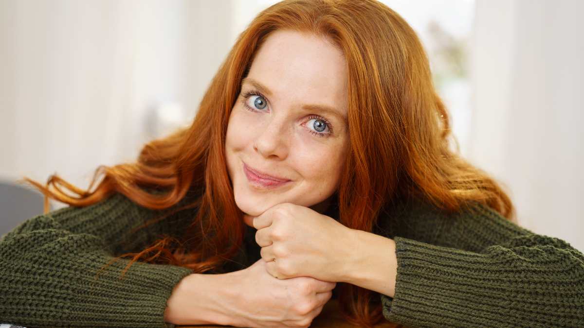 Pretty redhead woman with a cute grin leaning her chin on her hands as she leans on a wooden table.