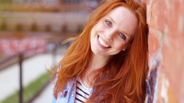 Pretty redhead woman with a lovely friendly smile leaning against a brick wall in an urban environment beaming at the camera, close up head and shoulders.