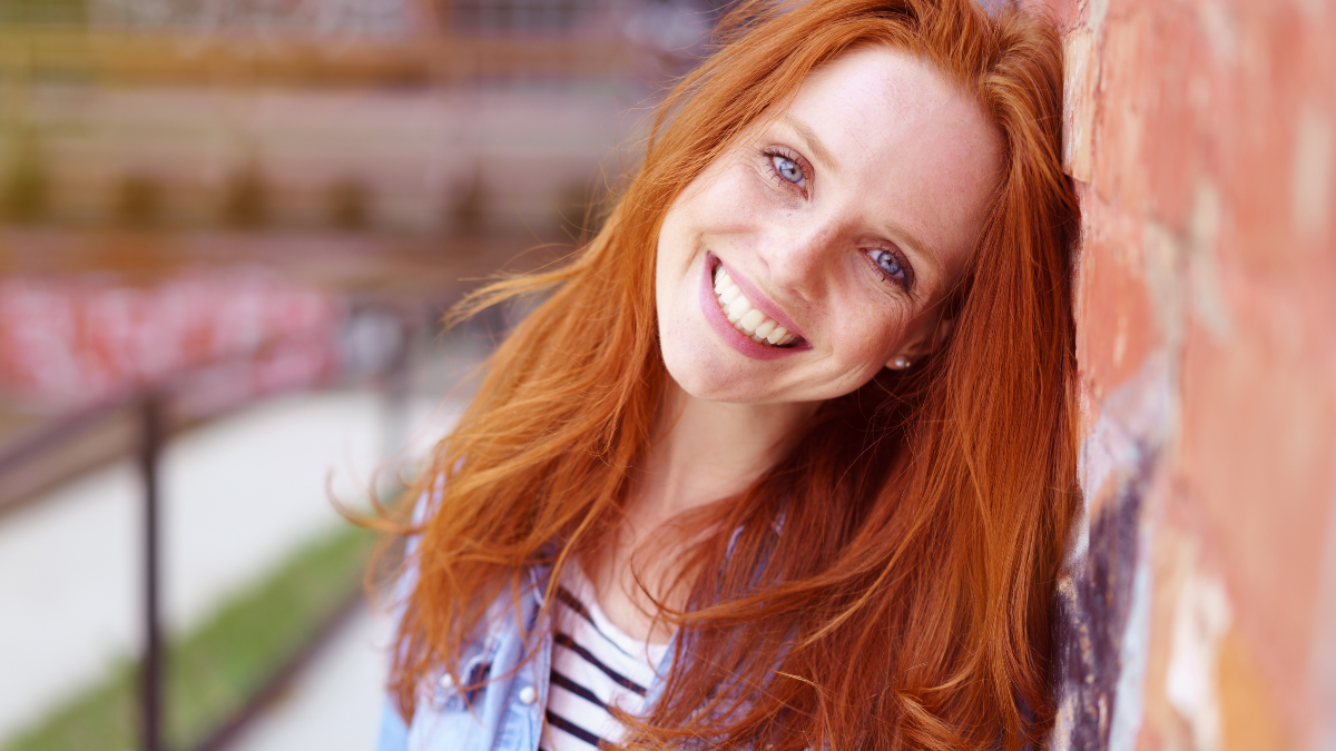 Pretty redhead woman with a lovely friendly smile leaning against a brick wall in an urban environment beaming at the camera, close up head and shoulders.