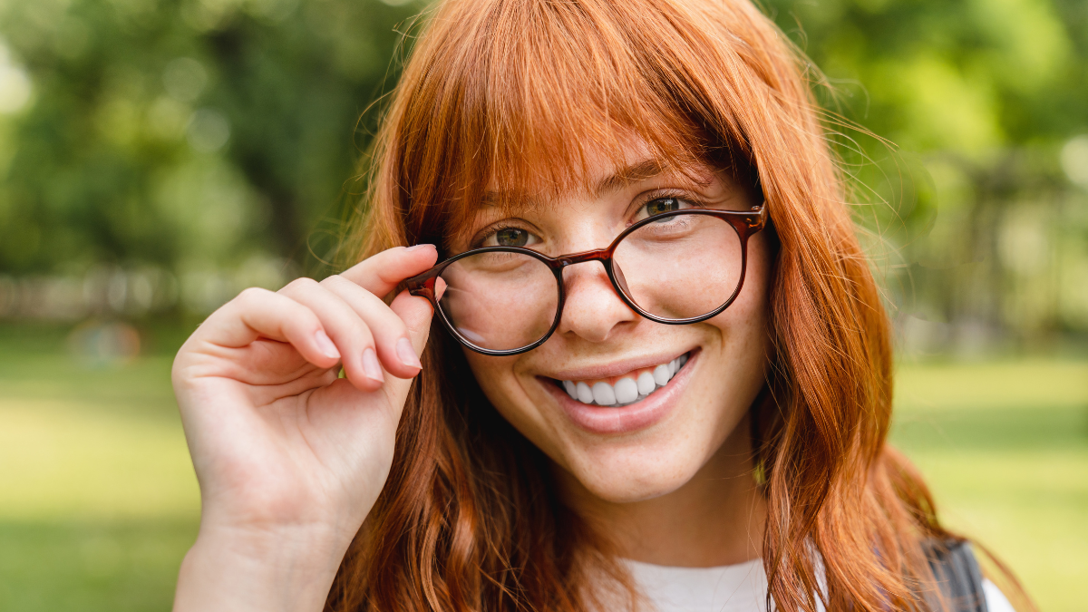 Beautiful smart caucasian young woman girl teenager smiling wearing glasses while looking at camera. Red-head schoolgirl walking in park forest.