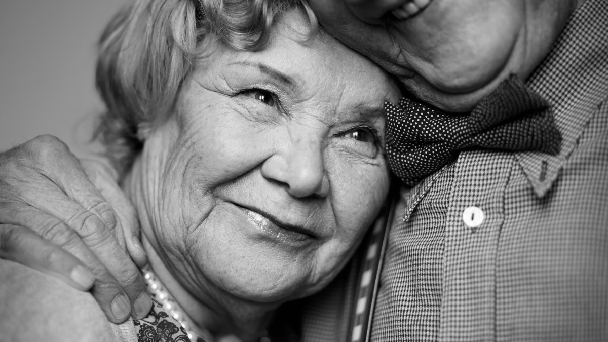 Black-and-white image of senior female being embraced by her husband.