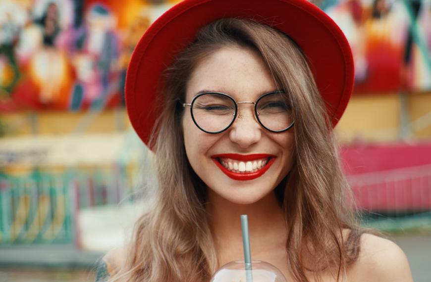 Cheerful young woman drinking summer refreshing lemonade, cocktail, drink. Red lips, shiny white teeth, hands, red hat, glasses, winking. Summer joyful lady looking to camera.