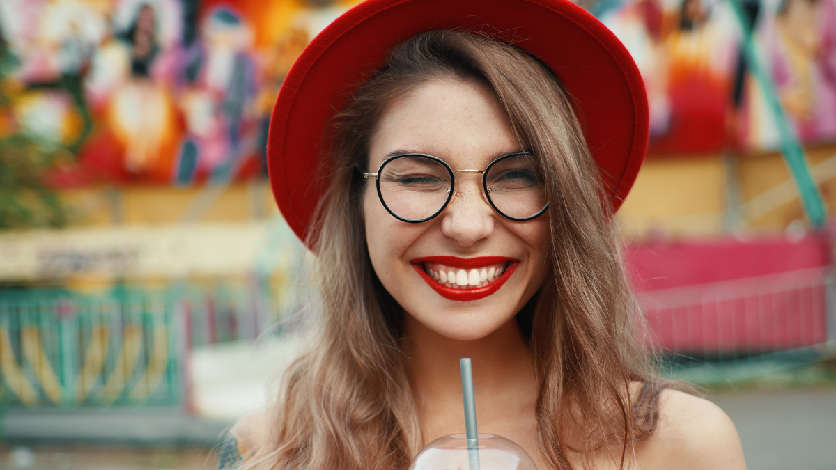 Cheerful young woman drinking summer refreshing lemonade, cocktail, drink. Red lips, shiny white teeth, hands, red hat, glasses, winking. Summer joyful lady looking to camera.