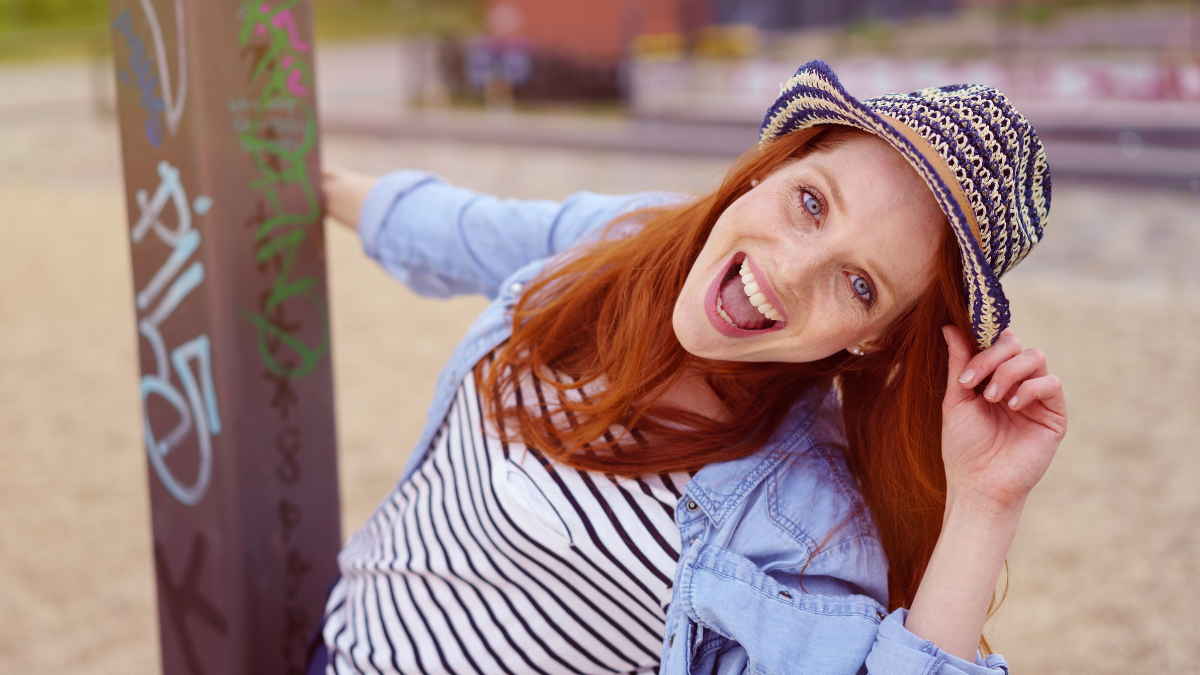 Single joyful young woman with red hair under festive fedora hat holding on to and swinging around a graffiti covered pole.