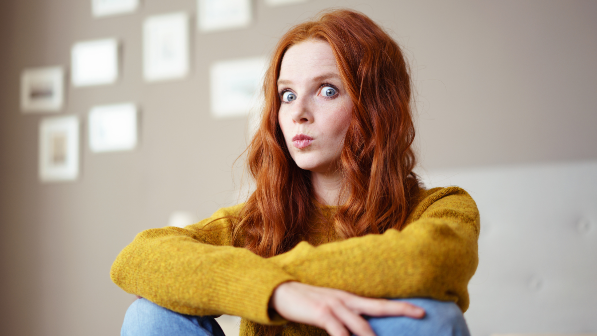 Pretty young redhead woman sitting on a bed looking suspiciously at the camera with wide eyes and a cute expression.
