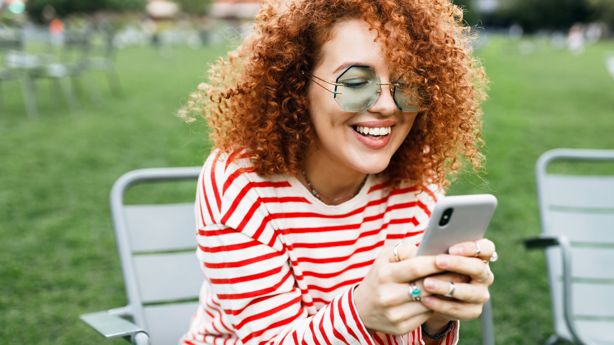 Outdoor portrait of hipster girl with red curly hair holding smartphone in hands sitting on chair in city park on lawn, using money-saving apps.