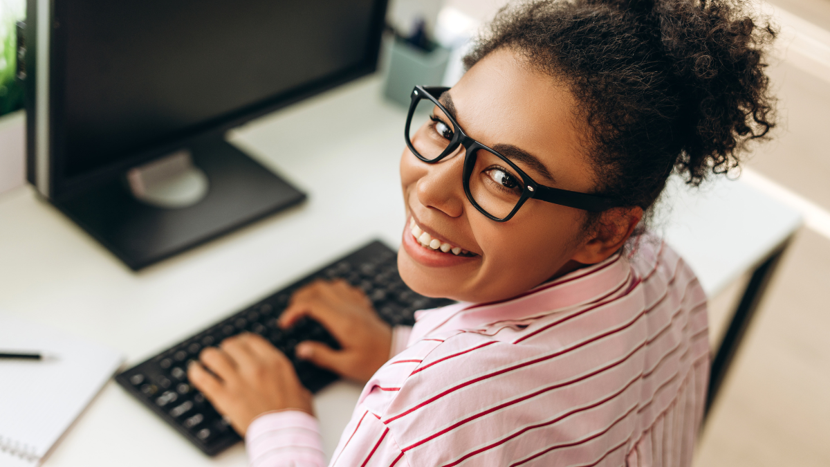 Cheerful young african american office worker wearing glasses and a pink shirt, sitting at her desk typing on a keyboard and looking over her shoulder, smiling.