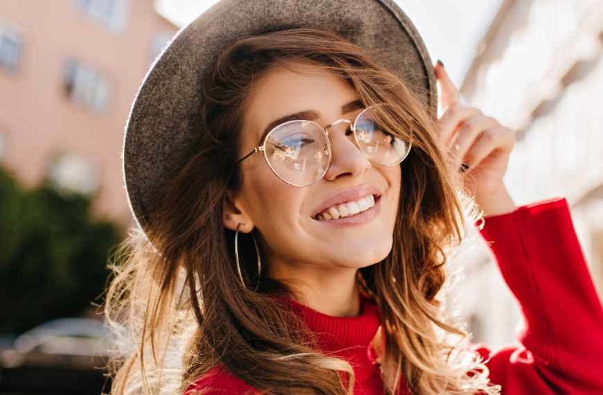 Close-up portrait of cheerful white woman in glasses touching her hat on blur background. Photo of fashionable girl with beautiful brown hair smiling to camera.