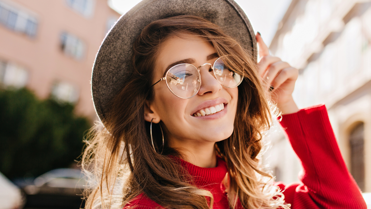 Close-up portrait of cheerful white woman in glasses touching her hat on blur background. Photo of fashionable girl with beautiful brown hair smiling to camera.