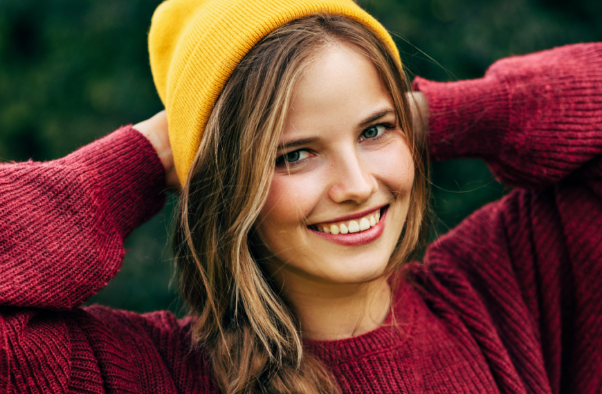 Portrait of a blonde young woman smiling broadly with a healthy toothy smile, wearing a red sweater, and yellow hat, posing on nature background in the park. People, travel, lifestyle.