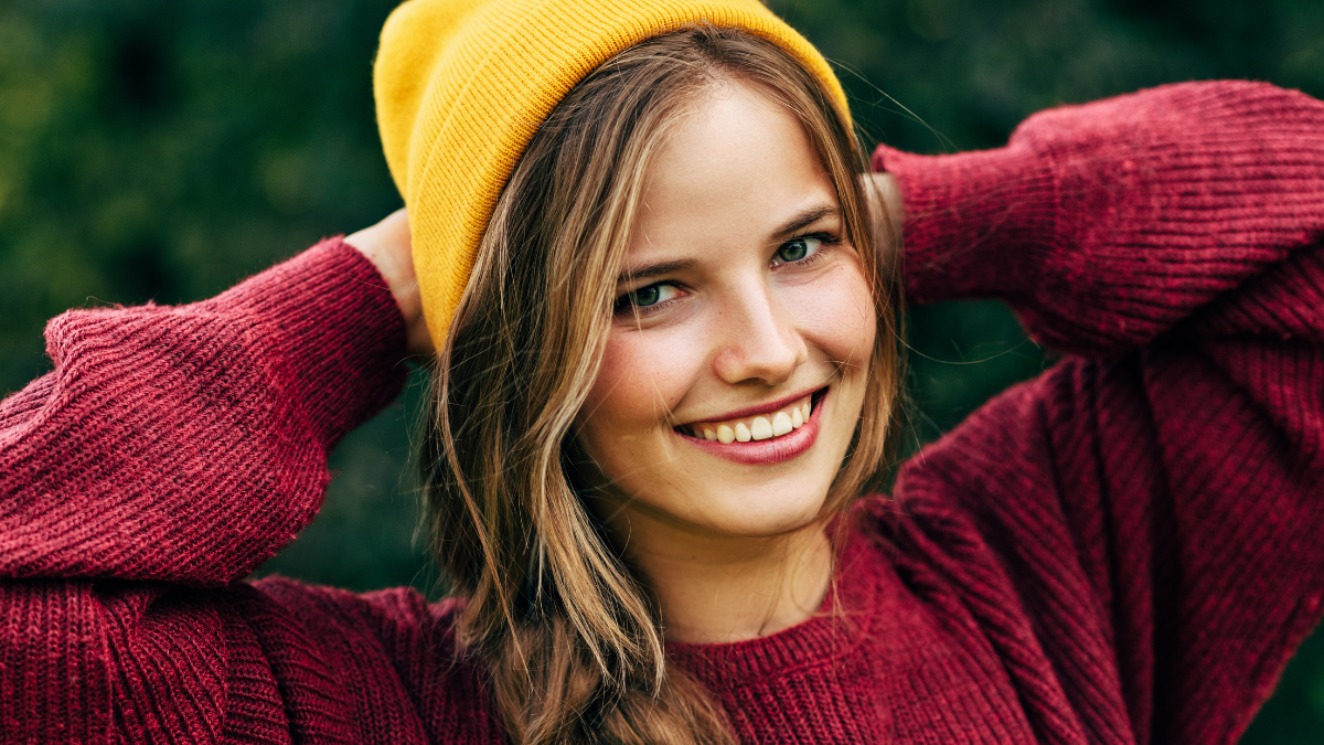 Portrait of a blonde young woman smiling broadly with a healthy toothy smile, wearing a red sweater, and yellow hat, posing on nature background in the park. People, travel, lifestyle.