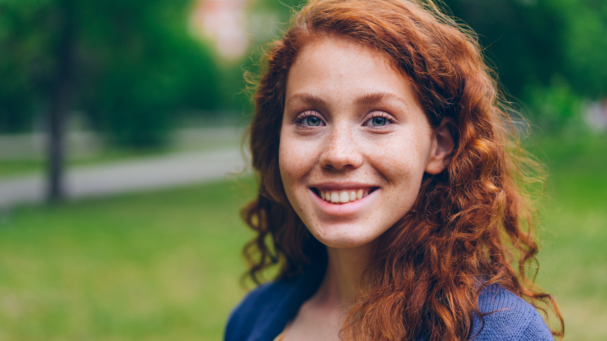 Close-up portrait of beautiful young lady with long red hair and freckles smiling looking at camera in park in summer. Nature, people and happiness concept.