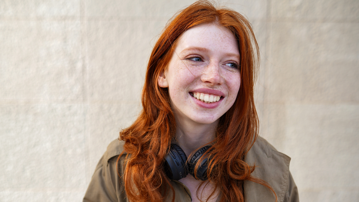 Happy teen stylish cool redhead fashion girl model standing on urban wall background laughing. Close up portrait of pretty joyful smiling teenage hipster girl with red hair looking away outdoors.