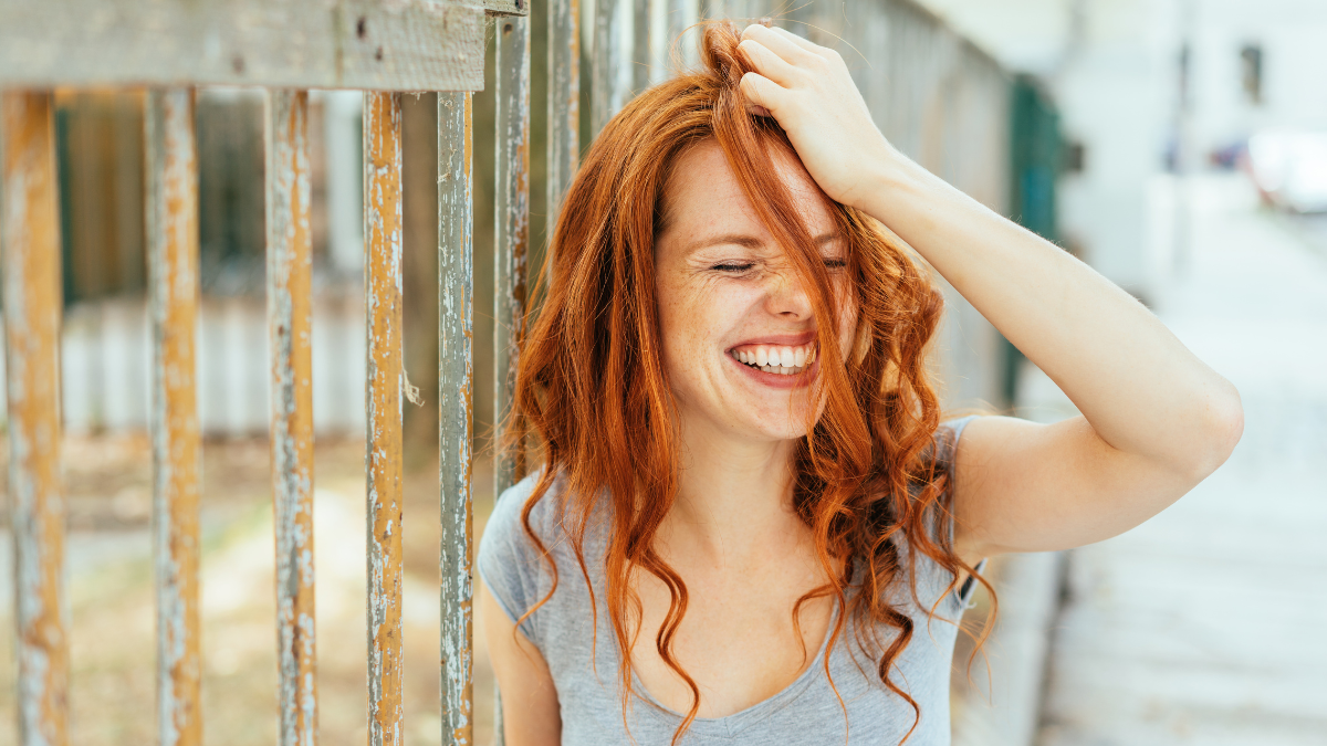 Grinning woman with red hair and grey shirt pulling her hair while leaning against tall wooden fence.