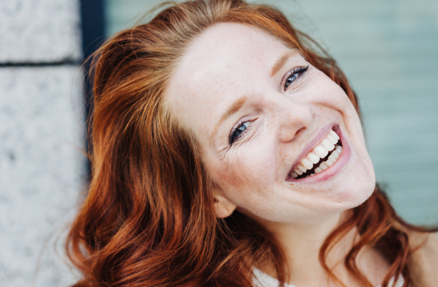 Happy young woman with a vivacious warm smile tilting her head to the side as she looks at the camera in a close up portrait outdoors.