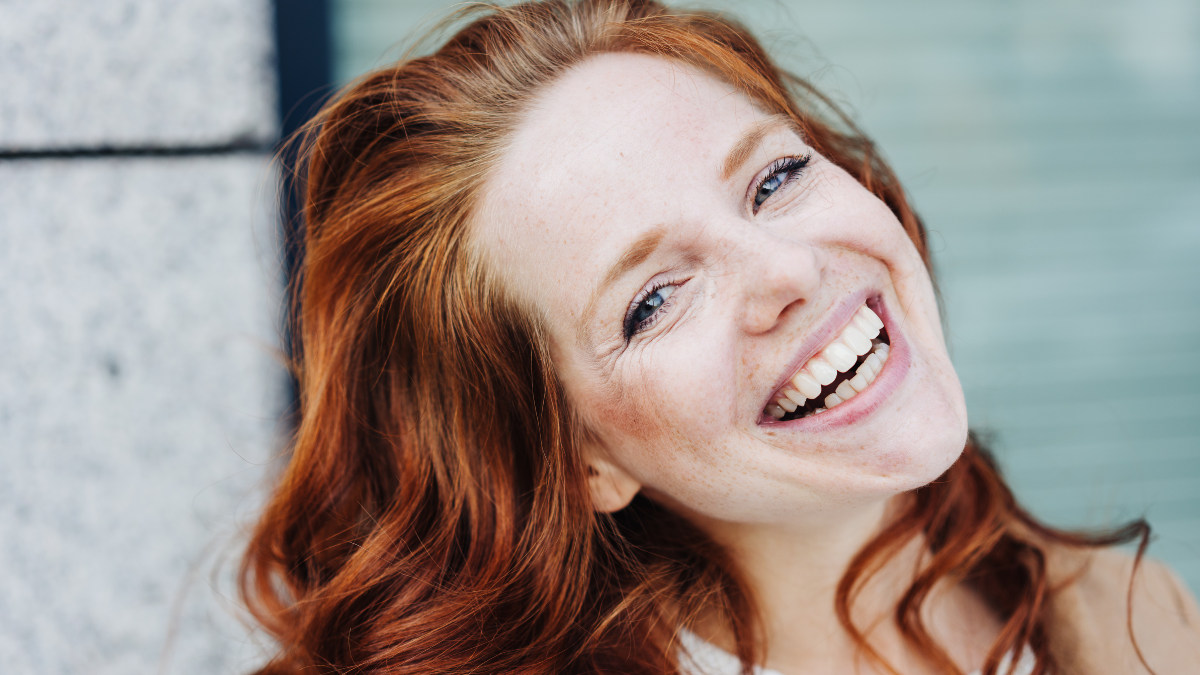 Happy young woman with a vivacious warm smile tilting her head to the side as she looks at the camera in a close up portrait outdoors.