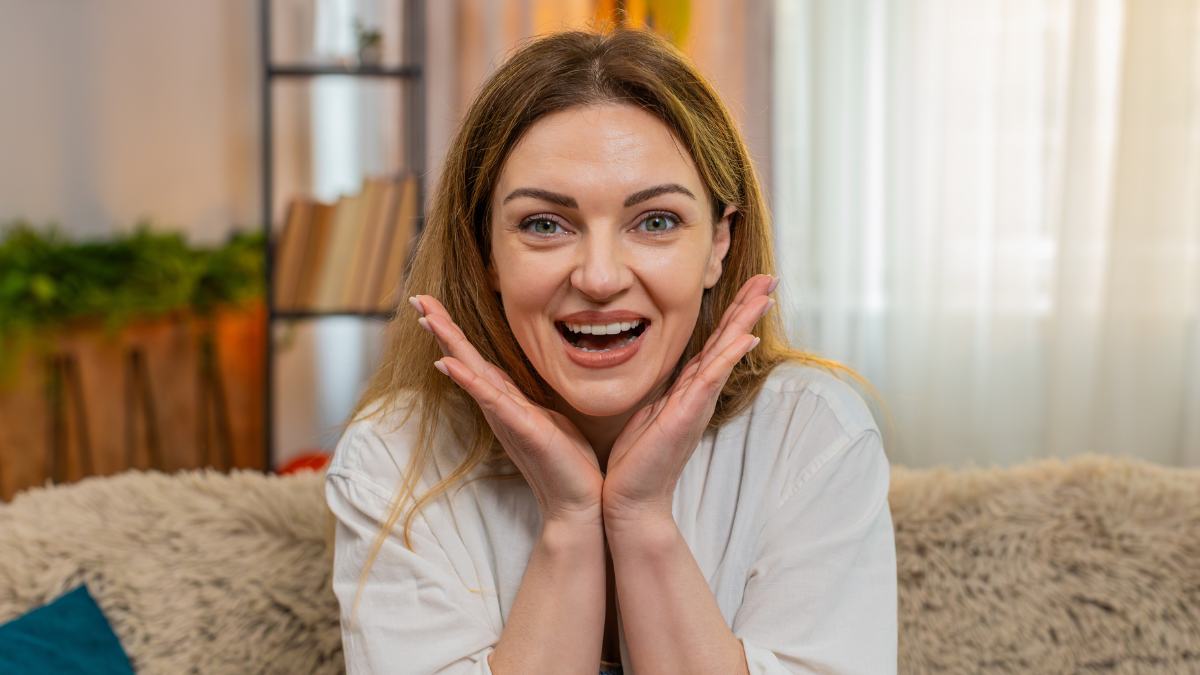 Middle-aged woman looks at camera on home sofa with surprise, saying wow and reacting with big emotion. Young adult girl shows amazement and visible shock, overwhelmed by unexpected moment or news.