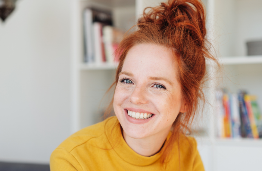 Close-up front portrait of a beautiful young woman in yellow with red hair, looking at camera with friendly face and laughing or smiling.