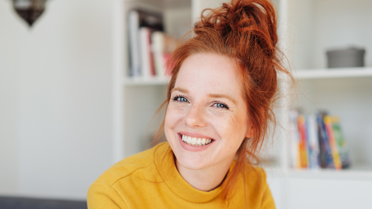 Close-up front portrait of a beautiful young woman in yellow with red hair, looking at camera with friendly face and laughing or smiling.