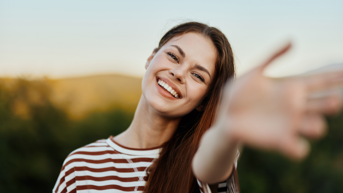 Woman smiling while looking at the camera and pulling her hands to the camera close-up in nature with a view of the mountains. Happy travel lifestyle follow me.