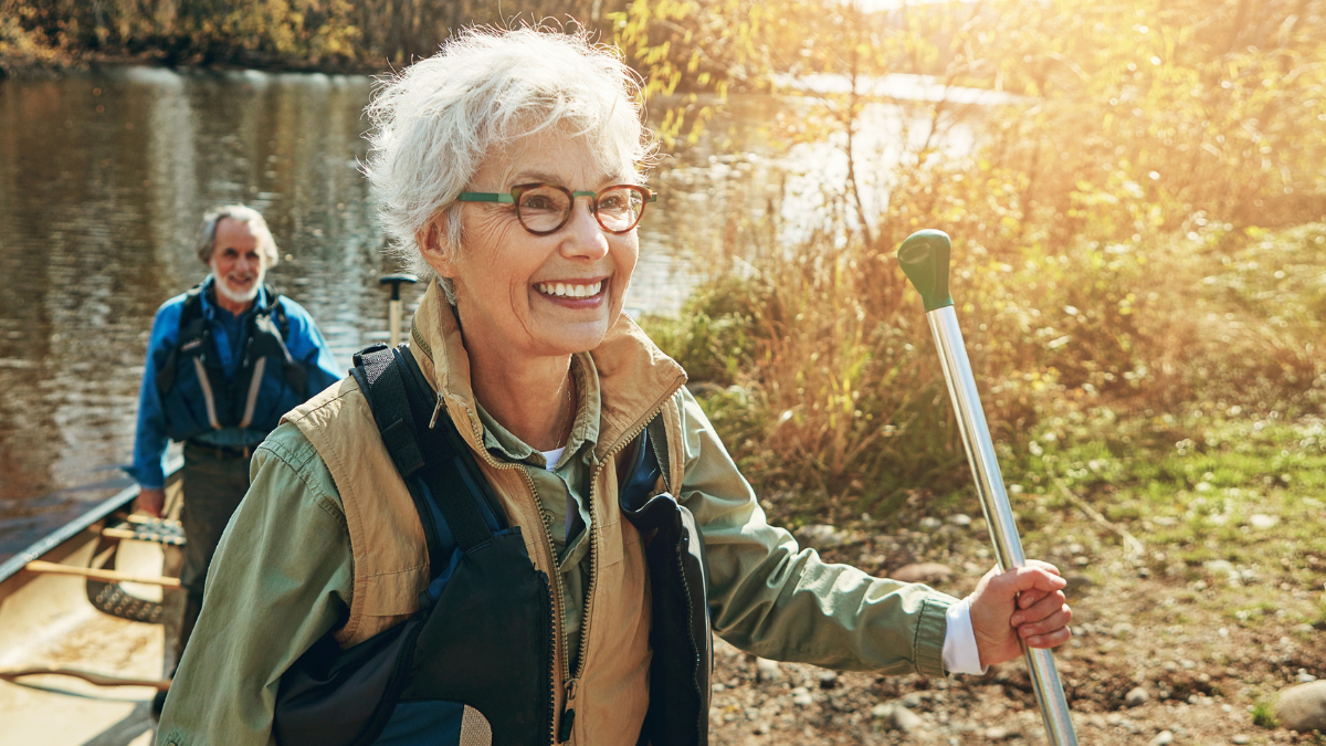 Senior woman, man and kayak adventure on lake in nature, freedom or travel for wellness. Happy couple, river and getting out of boat for walk in forest, vacation or holiday for retirement lifestyle.