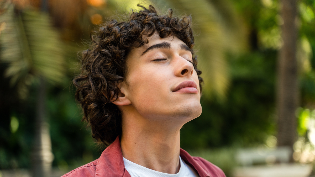 Tranquility. Portrait of a happy caucasian curly man relaxing with closed eyes while standing at the street at the park.