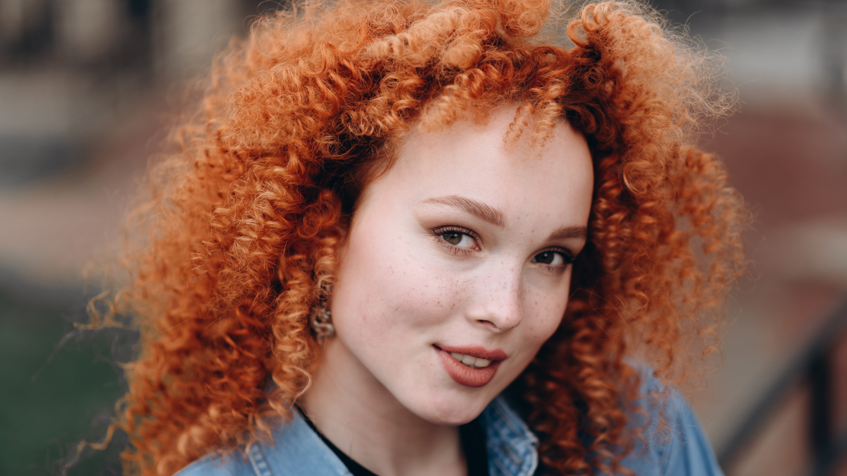 portrait of smiling young beautiful caucasian freckled girl with red curly hair in denim suit and black sweater in city street in sunny summer day, close-up view.