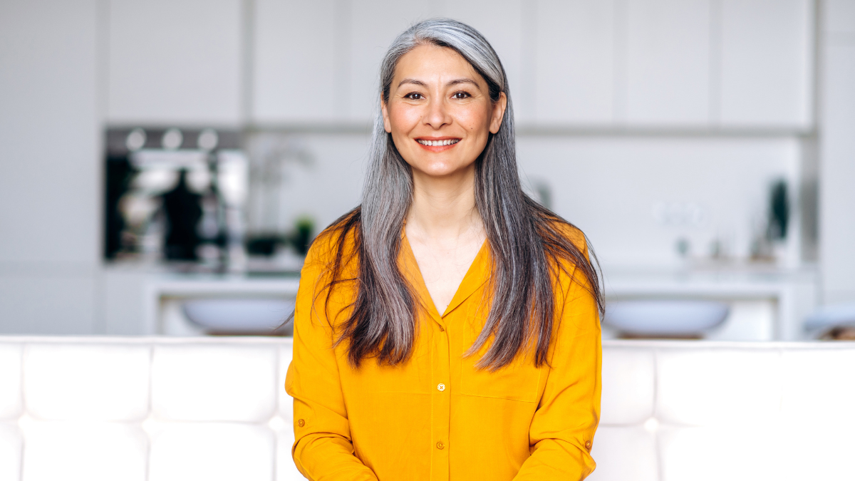 Portrait of a charming asian senior long gray hair lady wearing casual stylish clothes, sitting on couch at living room, looking directly at the camera with friendly smile.
