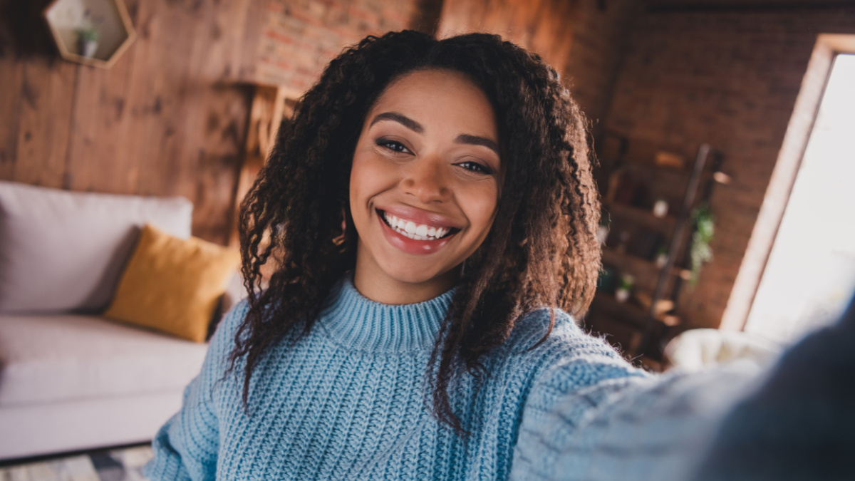 Charming young woman with afro hair smiling in a cozy living room during daylight, wearing a stylish knitted sweater.
