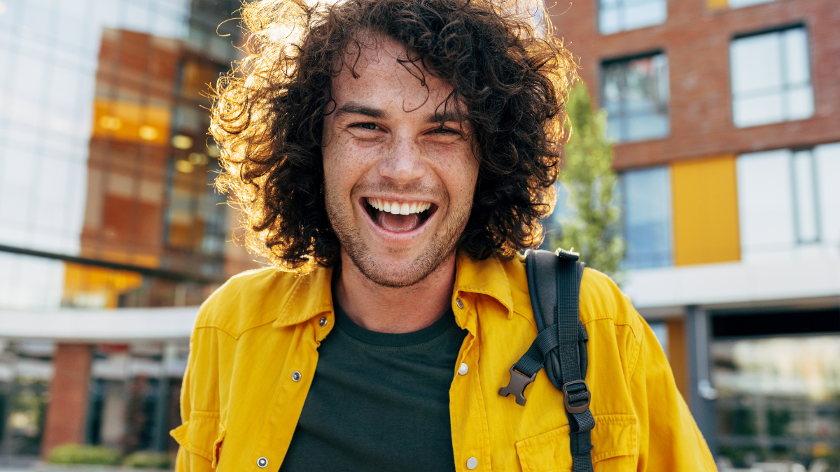 Outdoor portrait of cheerful man with curly hair, smiling broadly, posing for social advertisement in the city street. Happy student hipster male has joyful expression while walking outside.