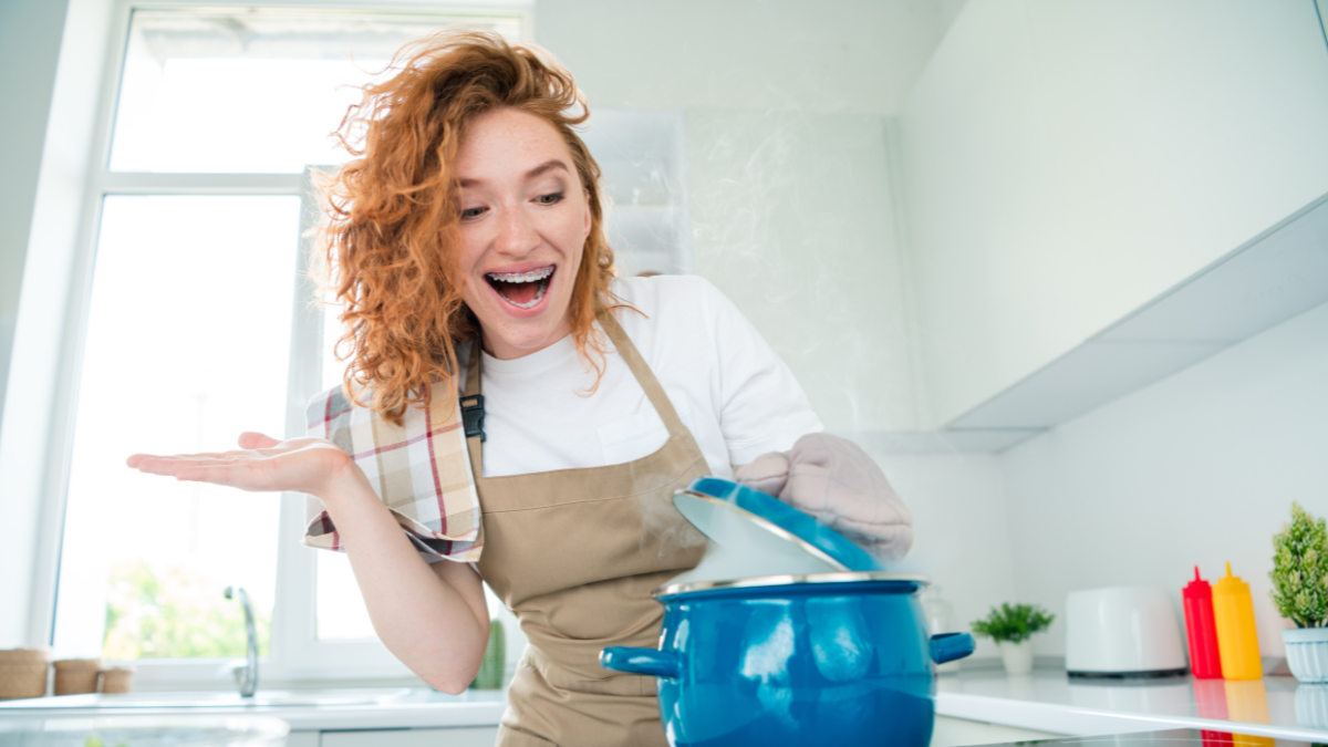 Charming redhead woman joyfully cooking a dish in cozy kitchen under natural daylight.