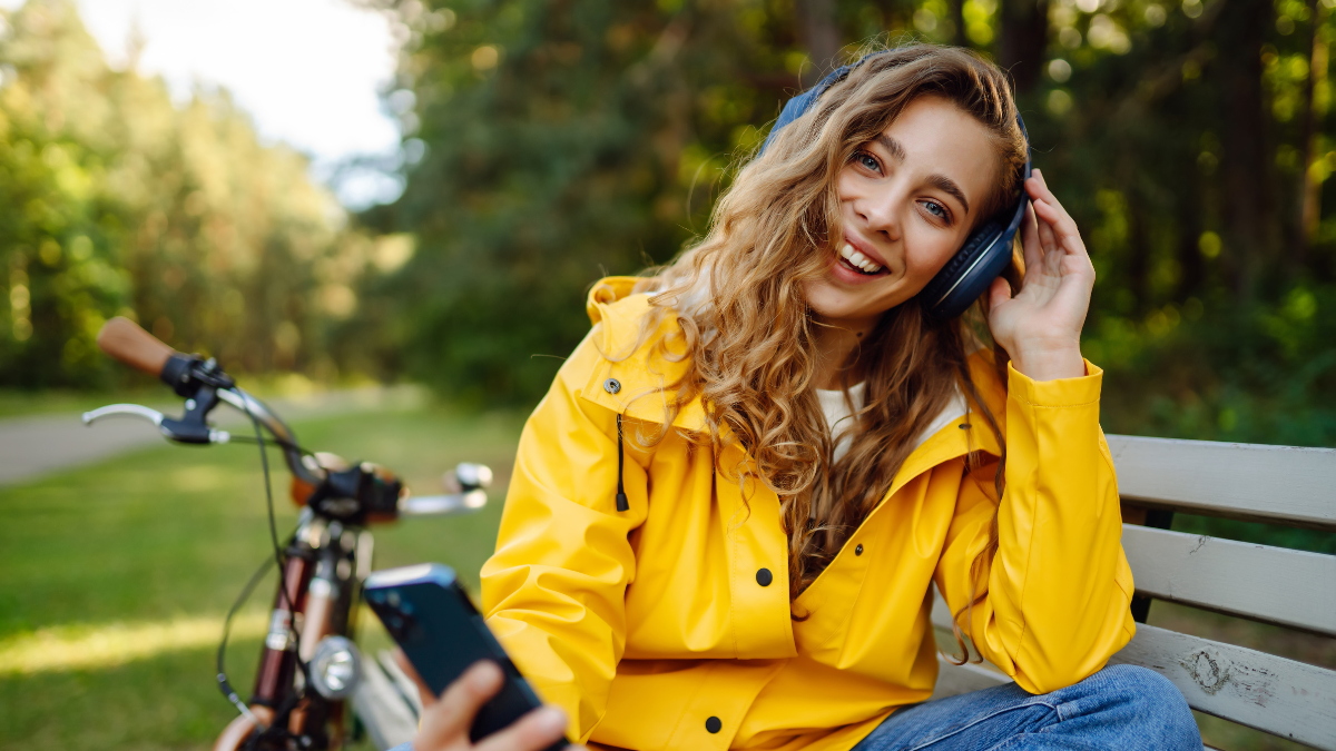 Happy young woman with headphones and smartphone rides a bicycle in a sunny park listening to music, podcast. Relax, nature concept. Lifestyle.