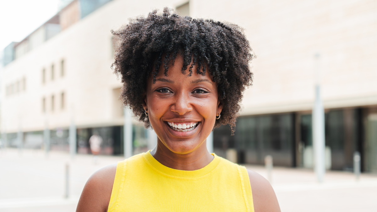 Close up individual portrait of one teenage african american girl smiling and looking at camera outside. A young adult woman staring front and laughing. Afro female gazing with friendly expression.