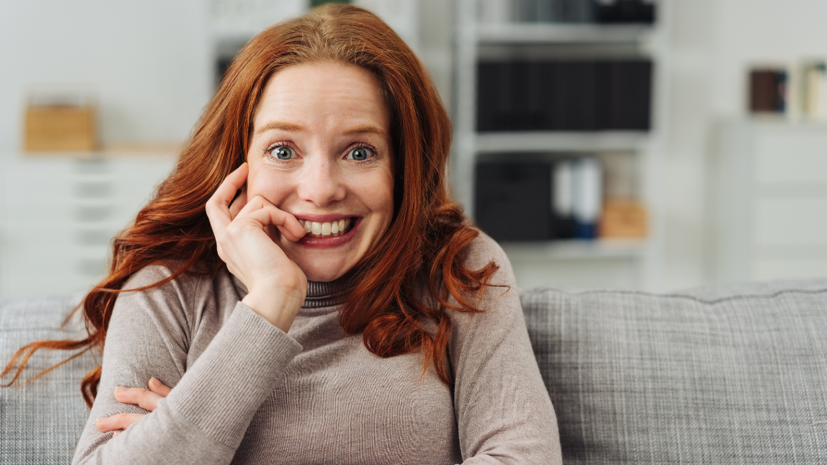 Young redhead woman with a look of excited anticipation staring at the camera with a big smile and wide eyes as she sits on a sofa at home.