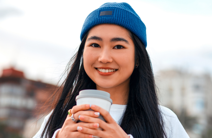 Close up portrait of a cute asian young woman wearing a hat drinking coffee or tea while standing outdoors.