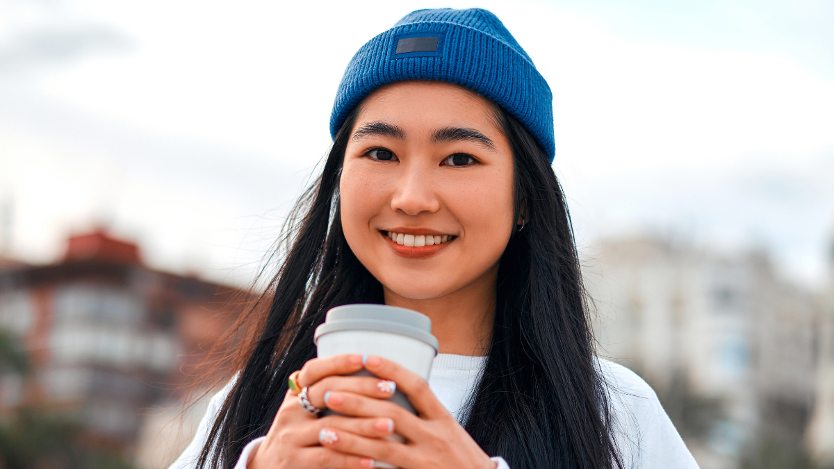 Close up portrait of a cute asian young woman wearing a hat drinking coffee or tea while standing outdoors.