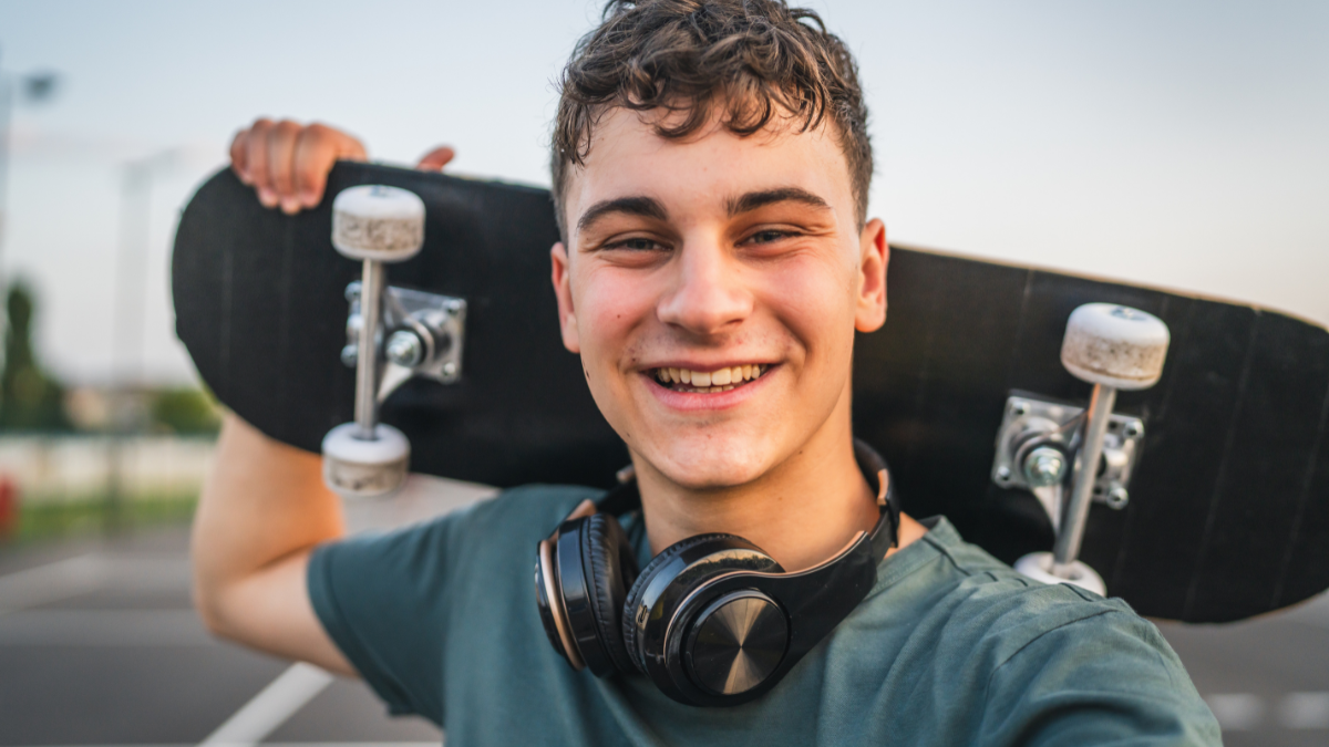 One man young adult caucasian teenager stand outdoor with skateboard on his shoulder and headphones posing at basketball court.