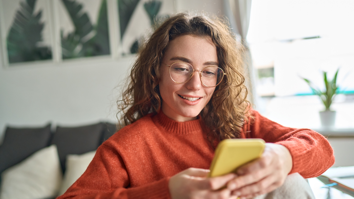Young smiling woman wearing glasses holding smartphone using cellphone modern technology, looking at mobile, checking cell phone apps, texting, browsing internet for shopping sitting at home.