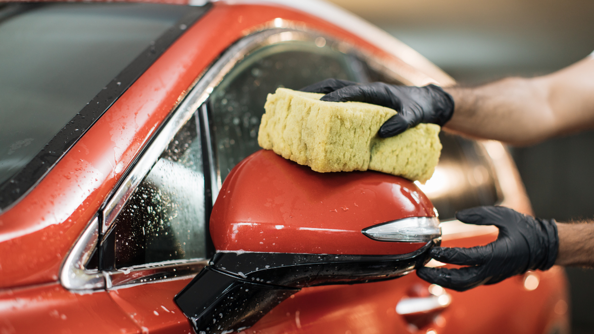 Cropped image of hand of car wash worker washing a soapy rearview mirror of red luxury car, holding sponge. Focus on the hand in black glove, holding sponge and washing car mirror. Cleaning, car wash.