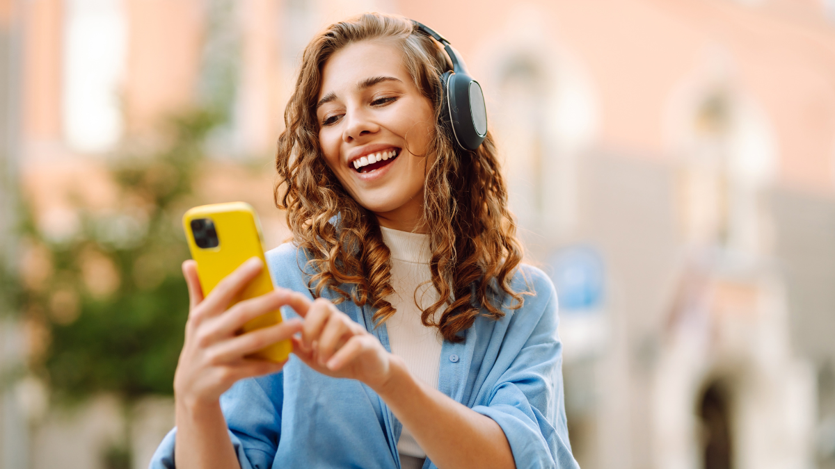 Happy relaxed woman in wireless headphones choosing, listening favorite music in urban city sunny street. Travel, tourism, technology, blogging concept.