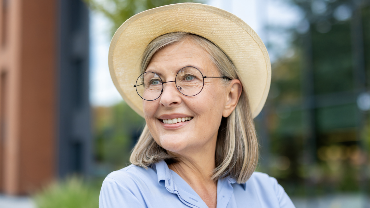 An elderly woman wearing glasses and a hat smiles cheerfully in an outdoor setting, showcasing a portrait of her.