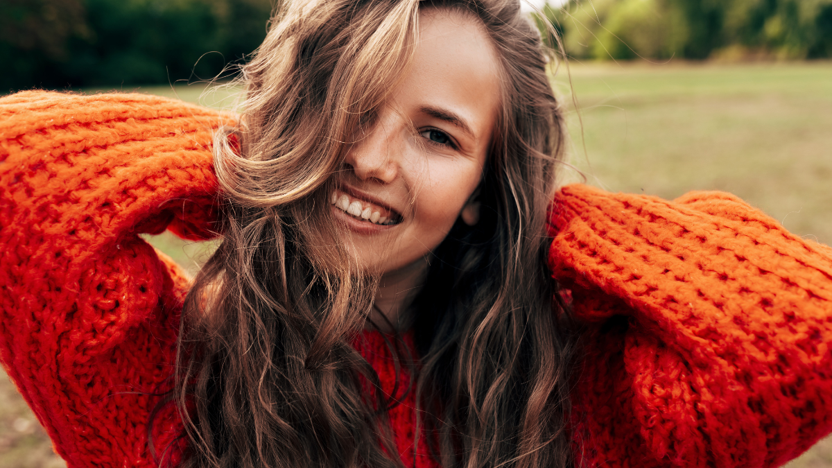 Outdoor portrait of a smiling young woman wearing a knitted orange sweater posing on nature background. The beautiful female has a joyful expression, resting in the park.